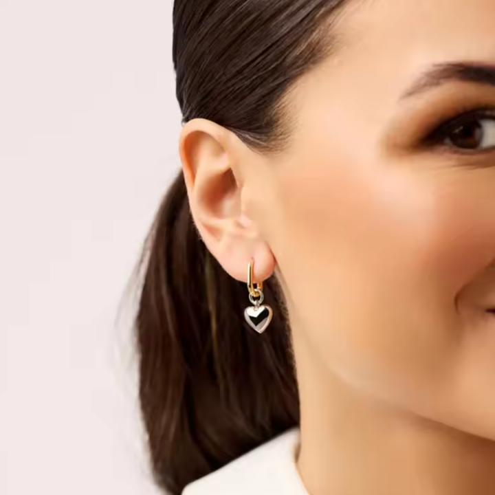 A close-up of a woman's ear wearing a small gold hoop earring with a dangling heart charm that has a black and white pattern, set against a clean light background.