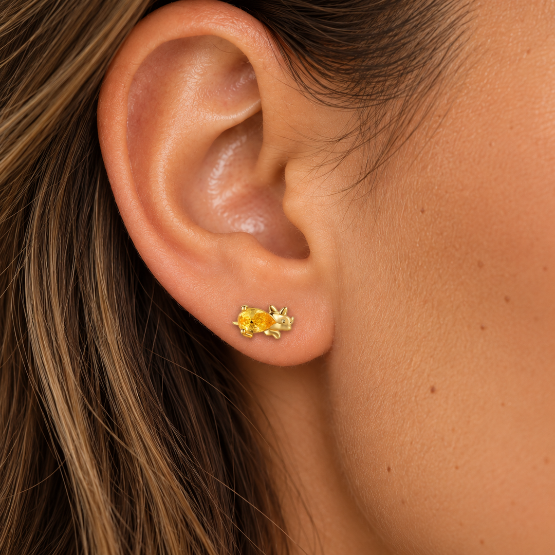 A close-up of a person's ear wearing a pair of small yellow gold stud earrings with clear gemstones in a prong setting.