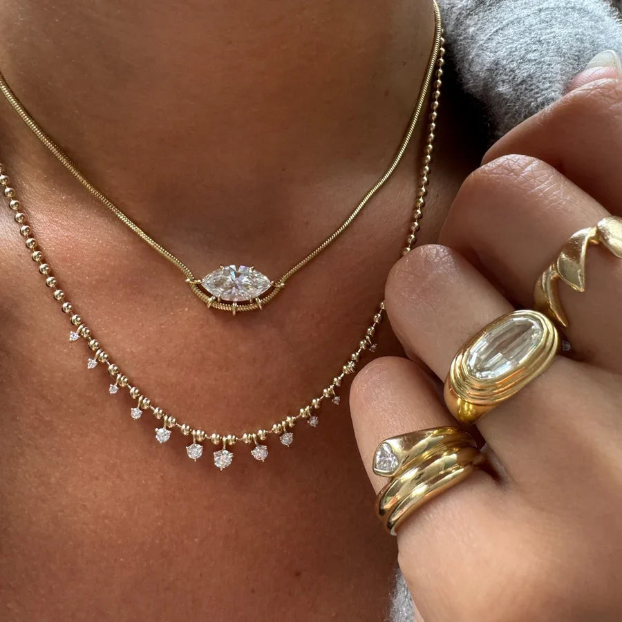 Gold layered necklaces worn on a model's neck, featuring a gold snake chain with an oval marquise-cut clear gemstone pendant and a delicate gold beaded chain with small heart-shaped clear gemstone drops. The model's hand is adorned with multiple gold rings.