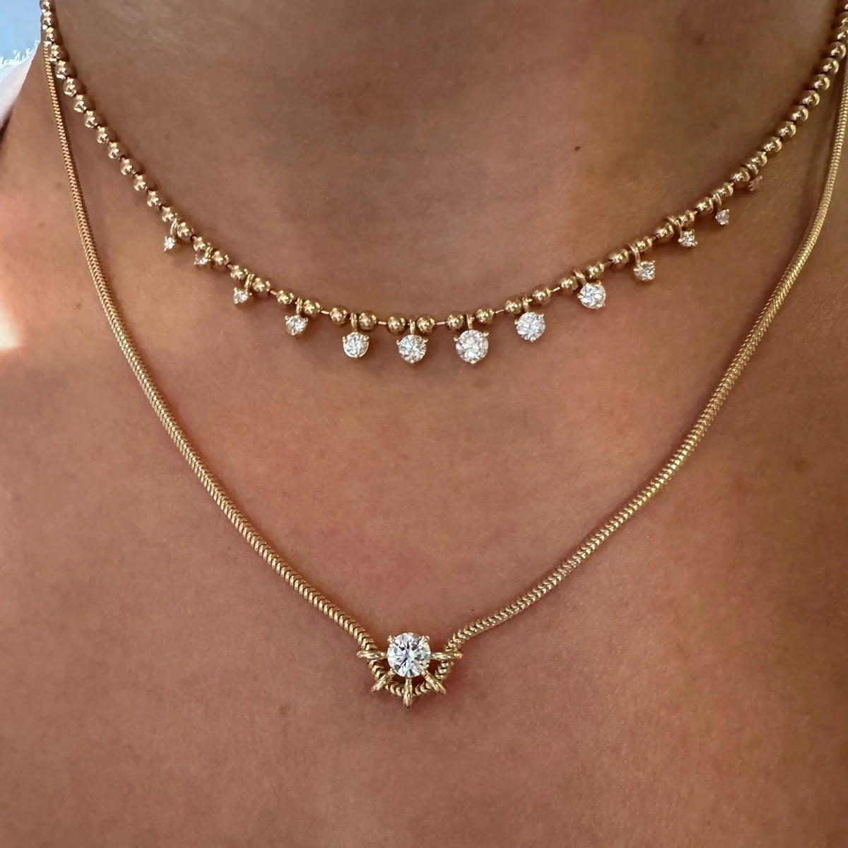 A close-up of a person's neck and upper chest wearing a layered gold necklace set. The top necklace has small gold beads and clear gemstone drops, while the longer chain necklace features a gold snake chain and a central sunburst-style pendant with a clear round gemstone.